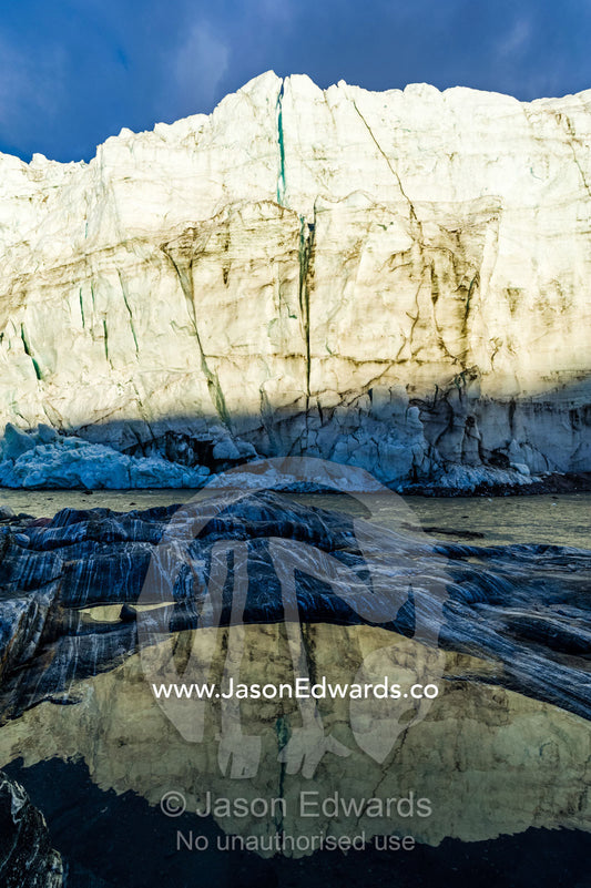 The setting sun reflects the sheer ice cliff of a glacier fracture zone in a pond. Russell Glacier, Greenland Ice Sheet, Qeqqata Municipality, Kangerlussuaq, Greenland.