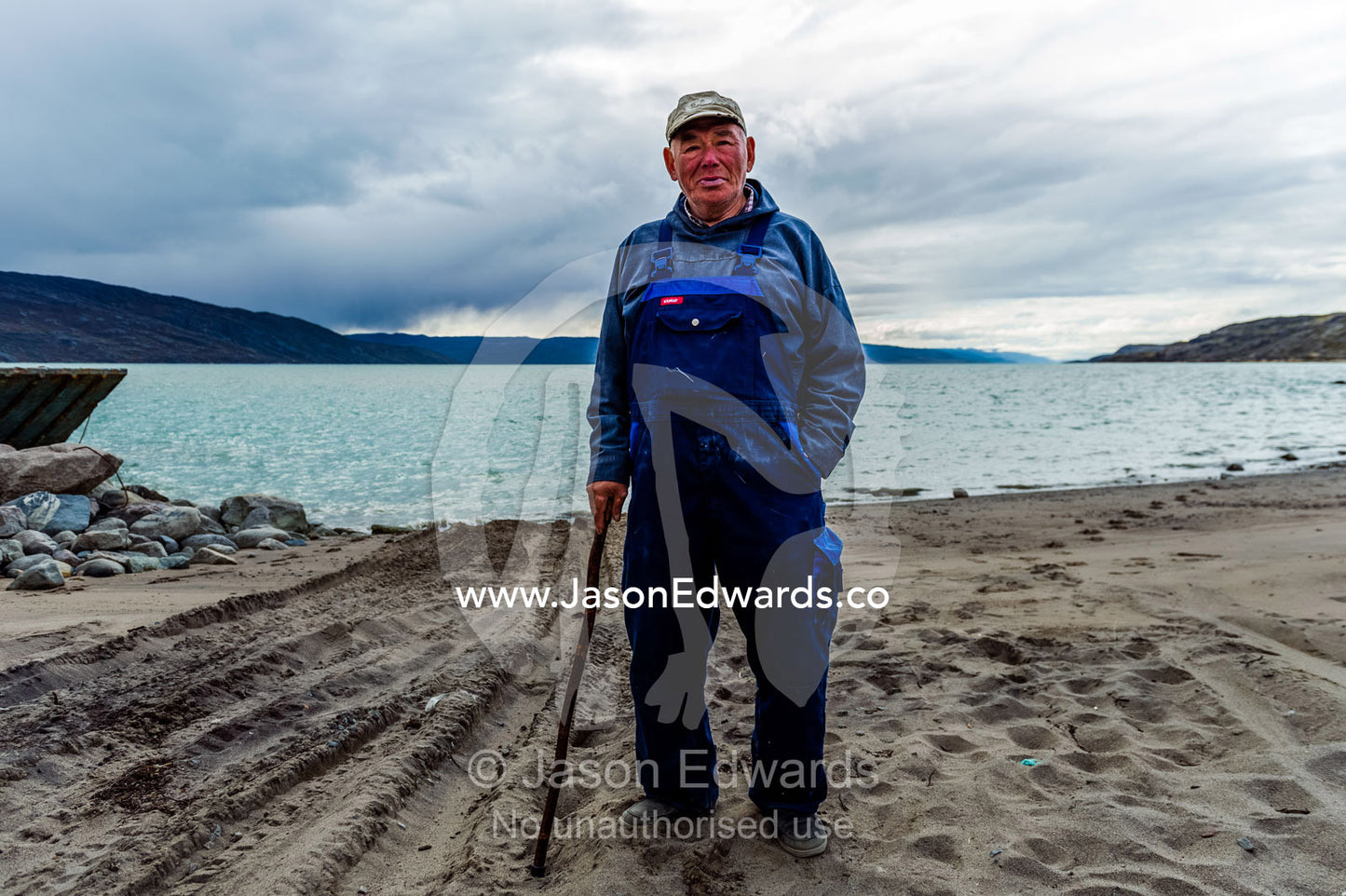 An elderly arctic fisherman standing on the shore of a fiord. Kangerlussuaq Port, Kangerlussuaq Fjord, Kangerlussuaq, Qeqqata Municipality, Greenland.