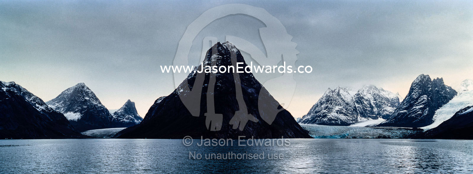 A cone-shaped mountain rises from the dark waters of an Arctic fjord beneath a storm laden sky. Fjord of Eternity, Maniitsoq, Sukkertoppen, Qeqqata Municipality, Greenland.