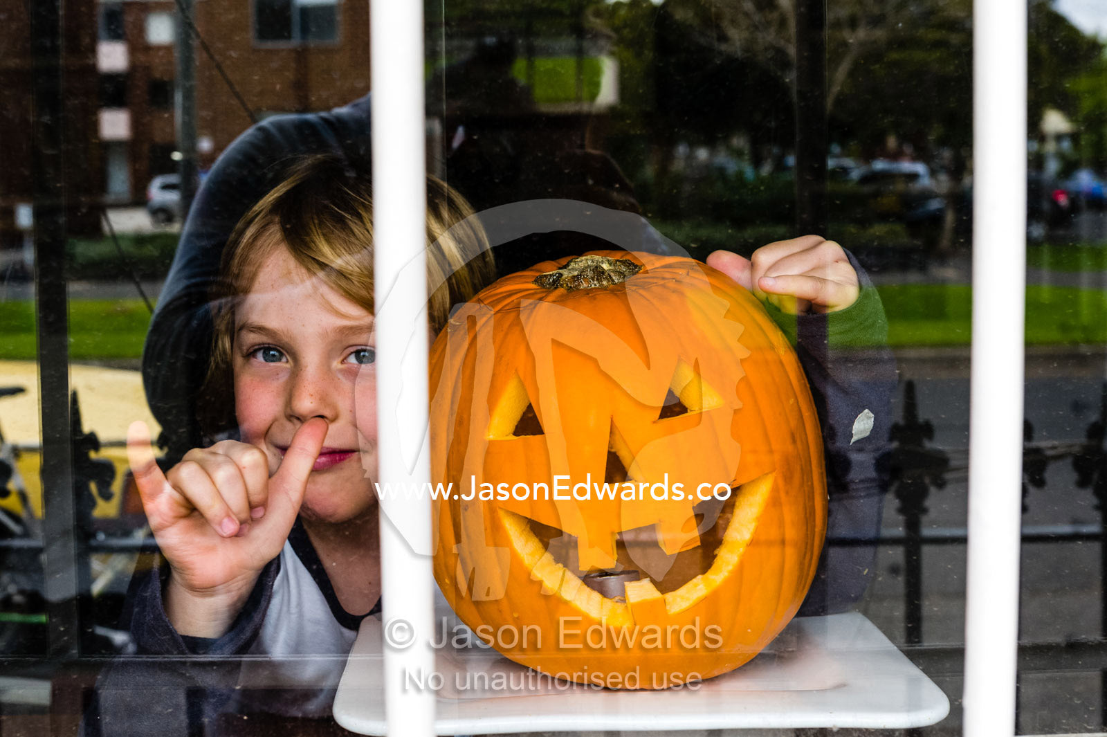 A boy smiles next to a grinning jack-o-lantern pumpkin in a house window on Halloween. Carlton North, Melbourne, Victoria, Australia.