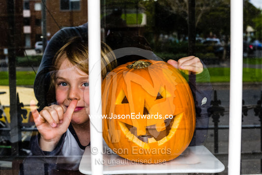 A boy smiles next to a grinning jack-o-lantern pumpkin in a house window on Halloween. Carlton North, Melbourne, Victoria, Australia.