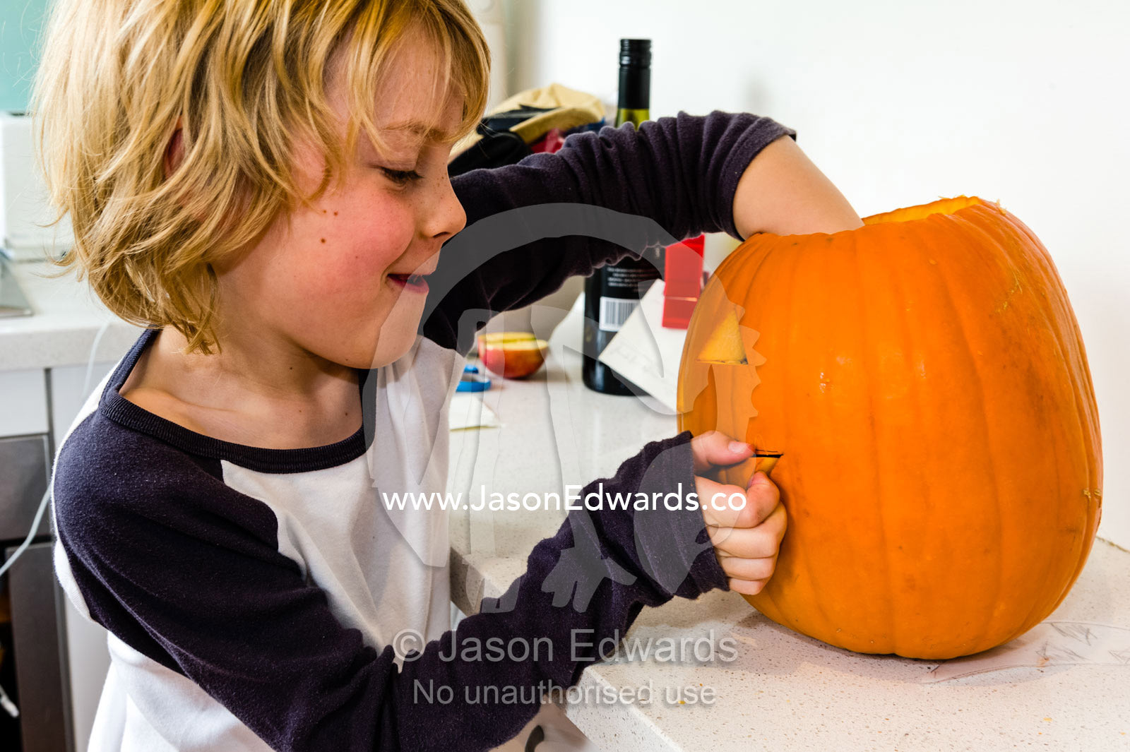 A boy gives a toothy smile whilst making a jack-o-lantern pumpkin in a kitchen for Halloween. Carlton North, Melbourne, Victoria, Australia.