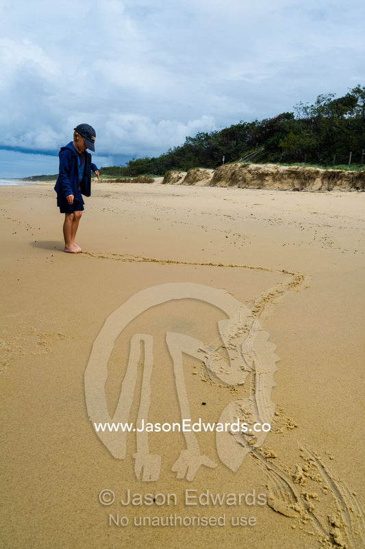 A boy draws patterns in the sand on a beach with his toes at low tide. Point Arkwright, Yaroomba Beach, Queensland, Australia.