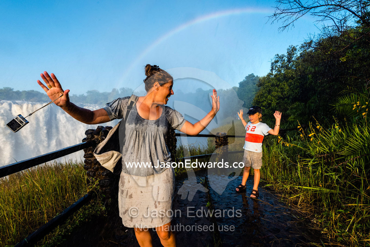 A mother and son dance beneath a rainbow in the rain falling from Victoria Falls. Zambezi River, Victoria Falls, Mosi-oa-Tunya National Park, Zambia.