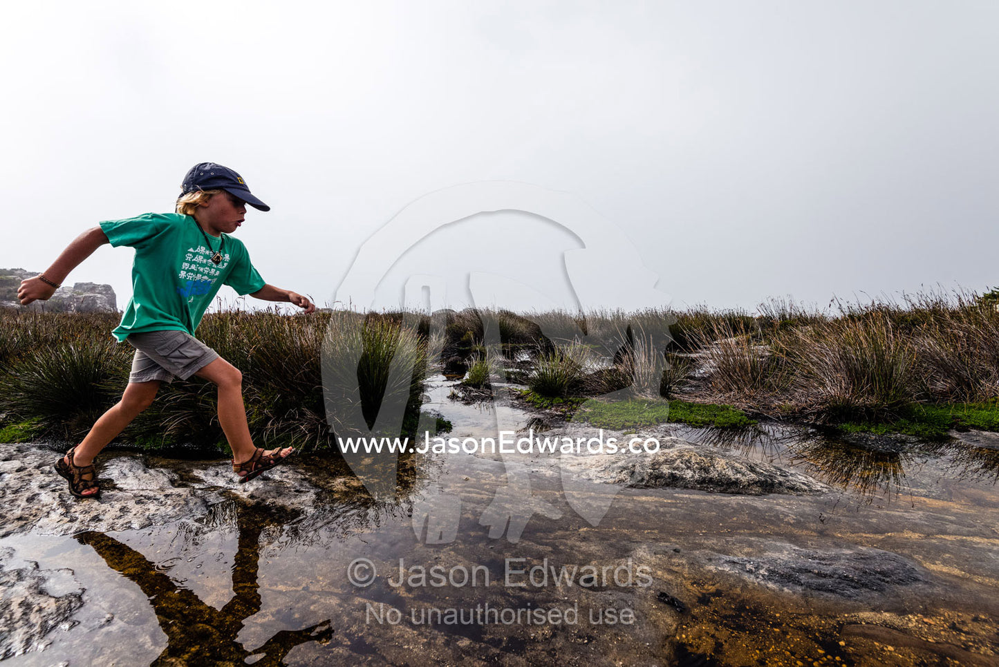 A boy leaps across pools of water captured in rock holes on the summit of Table Mountain. Table Mountain, Cape Town, Western Cape, South Africa.
