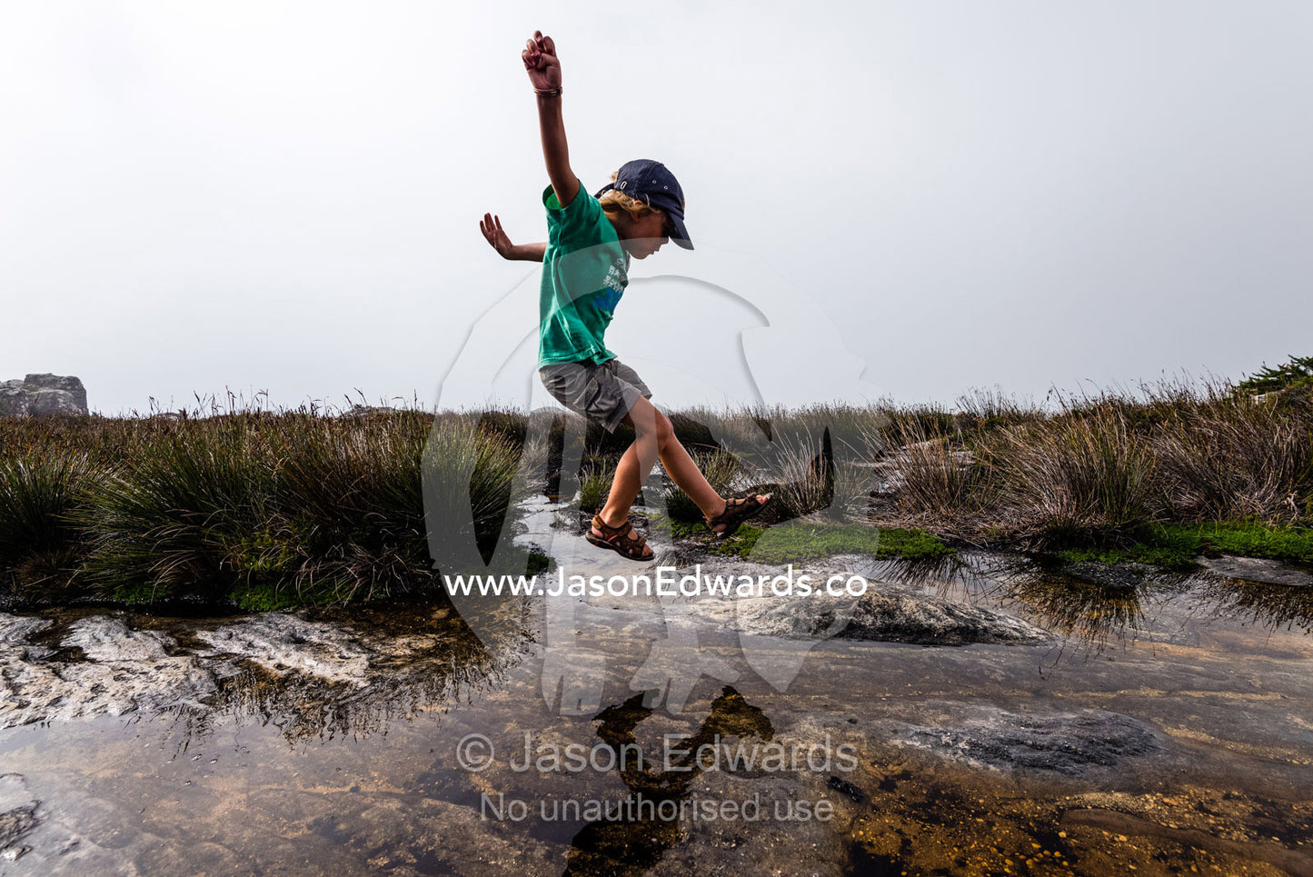 A boy leaps across pools of water captured in rock holes on the summit of Table Mountain. Table Mountain, Cape Town, Western Cape, South Africa.