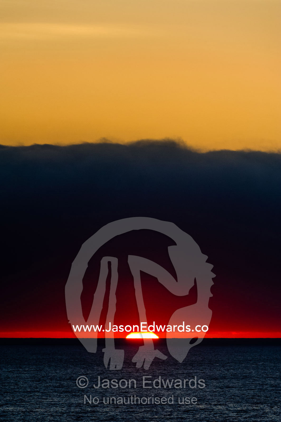 A slither of sunlight pierces a storm cloud above a darkened ocean at sunset. Cape Town, Table Bay, Western Cape, South Africa.