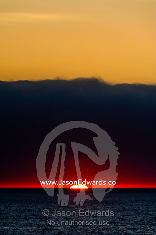 A slither of sunlight pierces a storm cloud above a darkened ocean at sunset. Cape Town, Table Bay, Western Cape, South Africa.