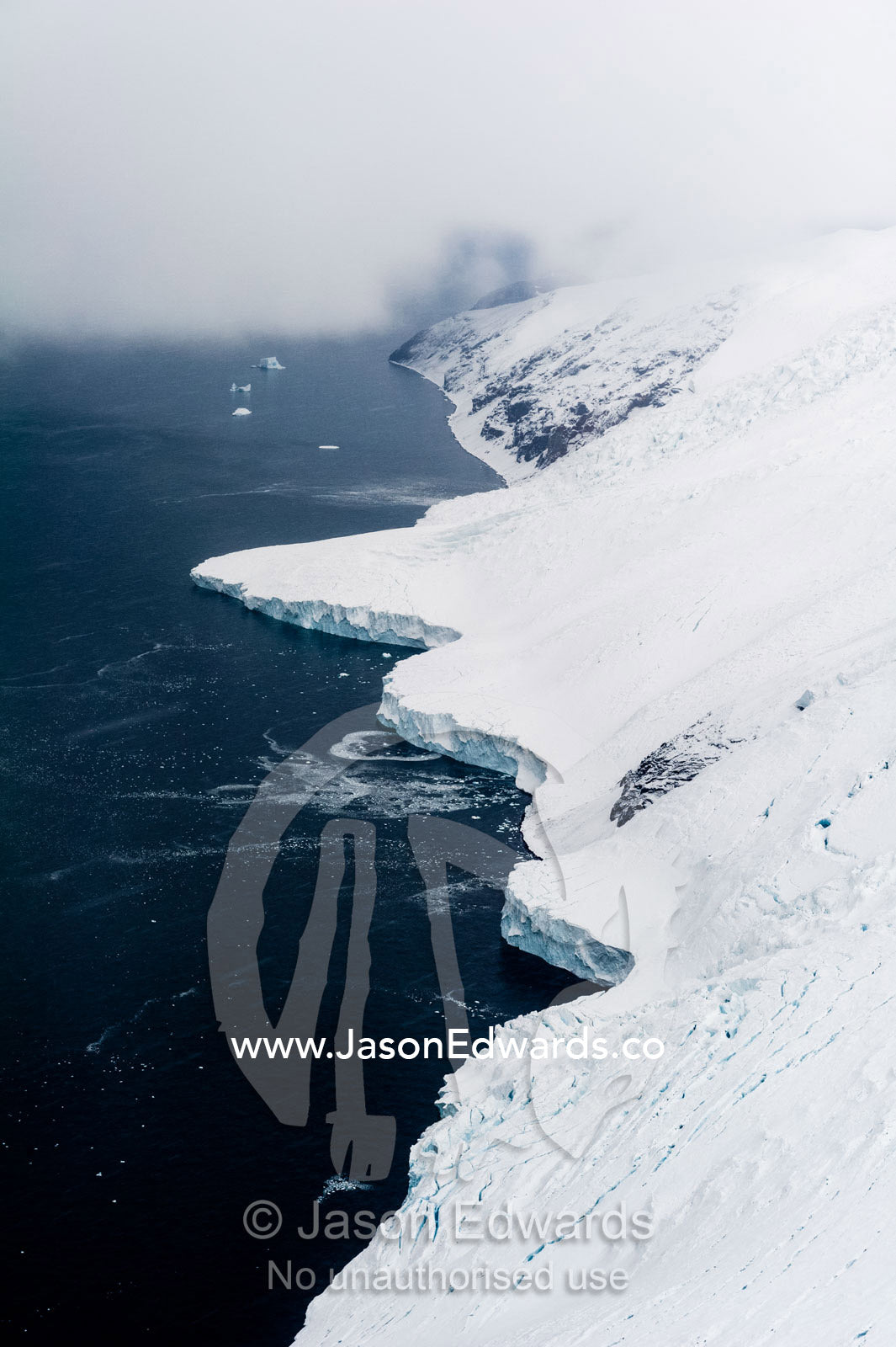Cliffs covered in snow and ice tower above a dark ocean surface. South West Face Mount Bird, Wohlschlag Bay, Ross Island, Antarctica.