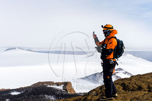 A mountain climber uses a cell phone to photograph Mount Erebus an active volcano from a mountaintop. Castle Rock, Hut Point Peninsula, Ross Island, Antarctica.