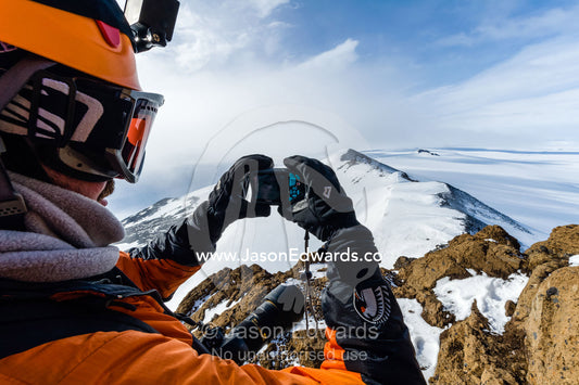 A mountain climber uses a pocket camera to photograph Mount Erebus an active volcano from a mountaintop. Breached Cone, Castle Rock, Hut Point Peninsula, Ross Island, Antarctica.