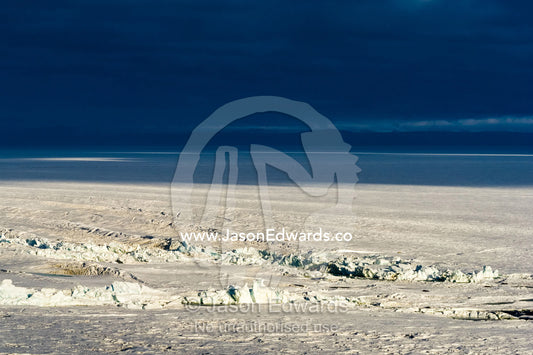 A sunray pierces a dark storm cloud illuminating jagged pressure ridges in the sea ice. Pram Point, McMurdo Sound, Ross Island, Antarctica.