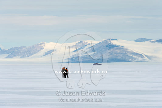 Researchers explore the vast frozen sea ice with a Coast Guard icebreaker in the distance. Turtle Rock, Erebus Bay, McMurdo Sound, Ross Island, Antarctica.