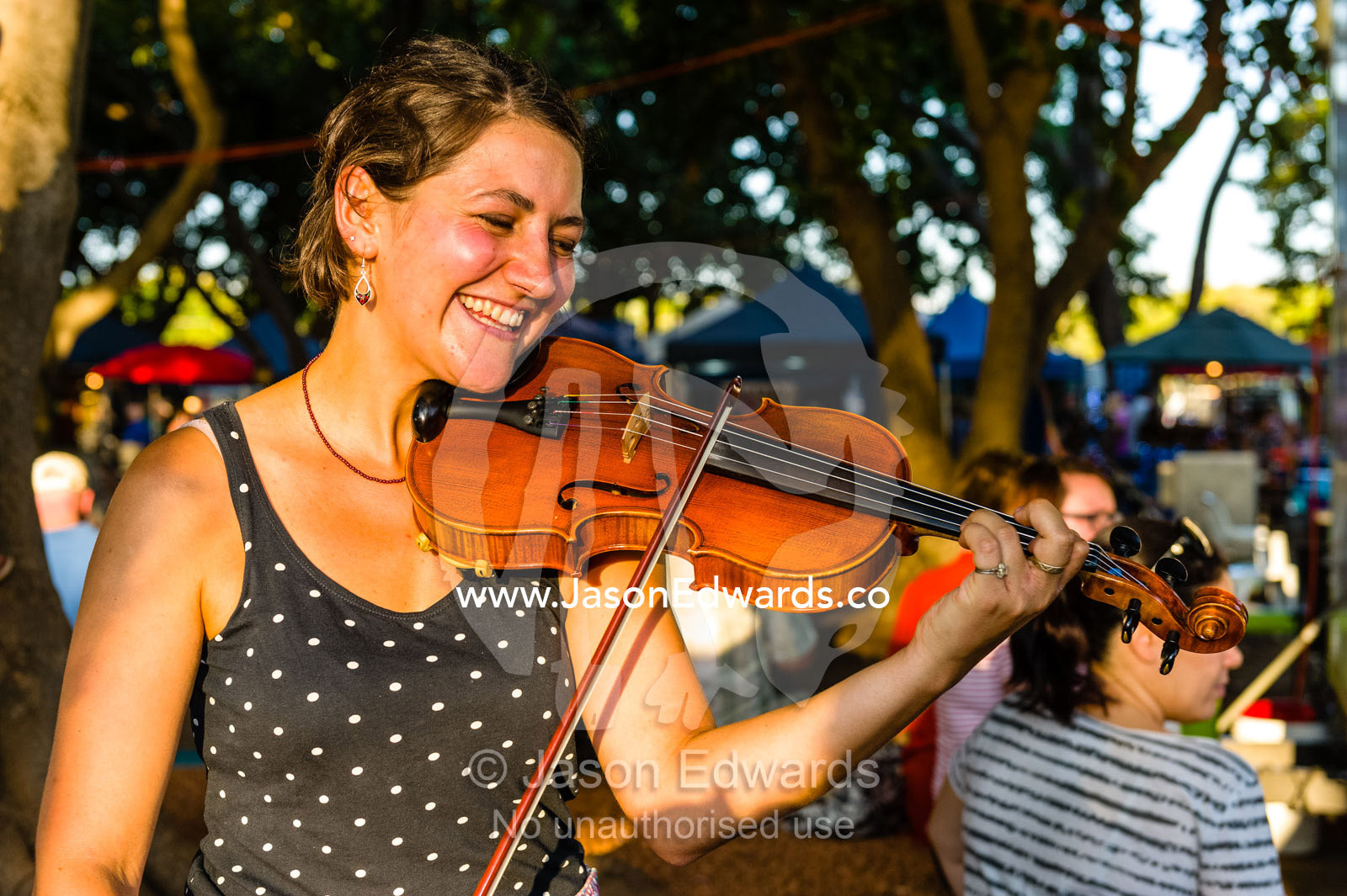 A street performer plays the fiddle for visitors at an open-air market. Mindil Beach Sunset Markets, Darwin, Northern Territory, Australia.