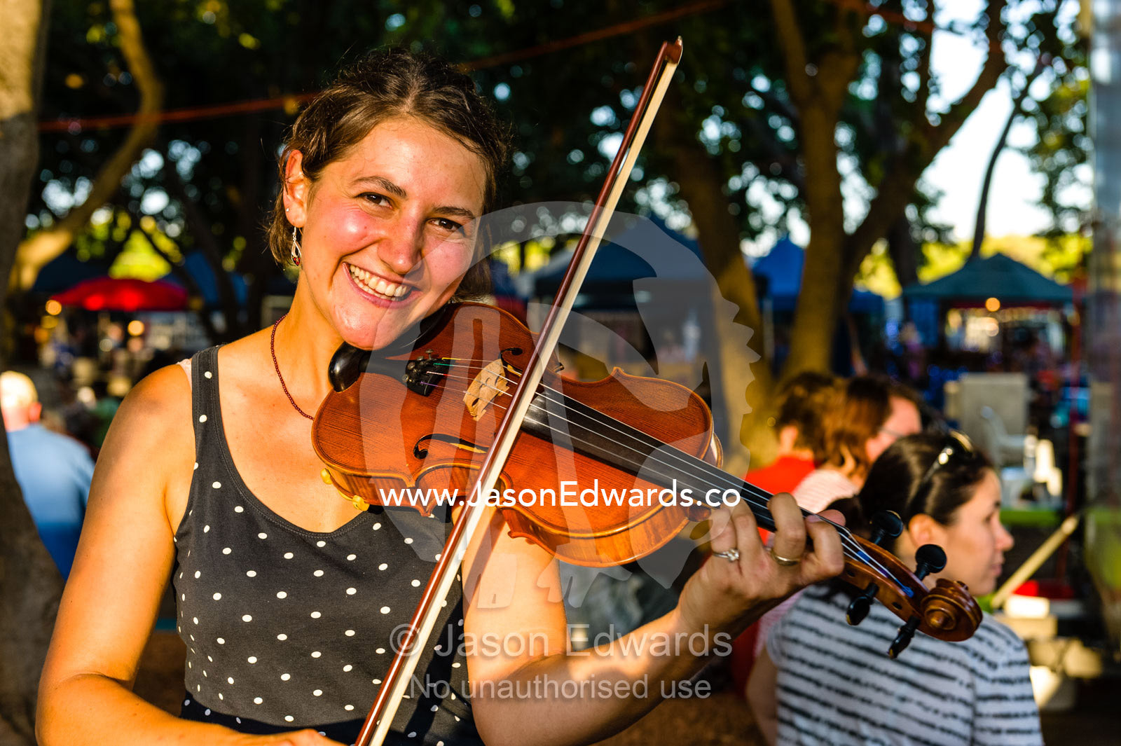 A street performer plays the fiddle for visitors at an open-air market. Mindil Beach Sunset Markets, Darwin, Northern Territory, Australia.