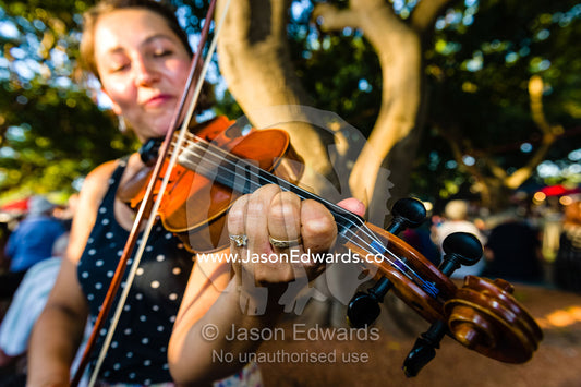 A street performer plays the fiddle for visitors at an open-air market. Mindil Beach Sunset Markets, Darwin, Northern Territory, Australia.