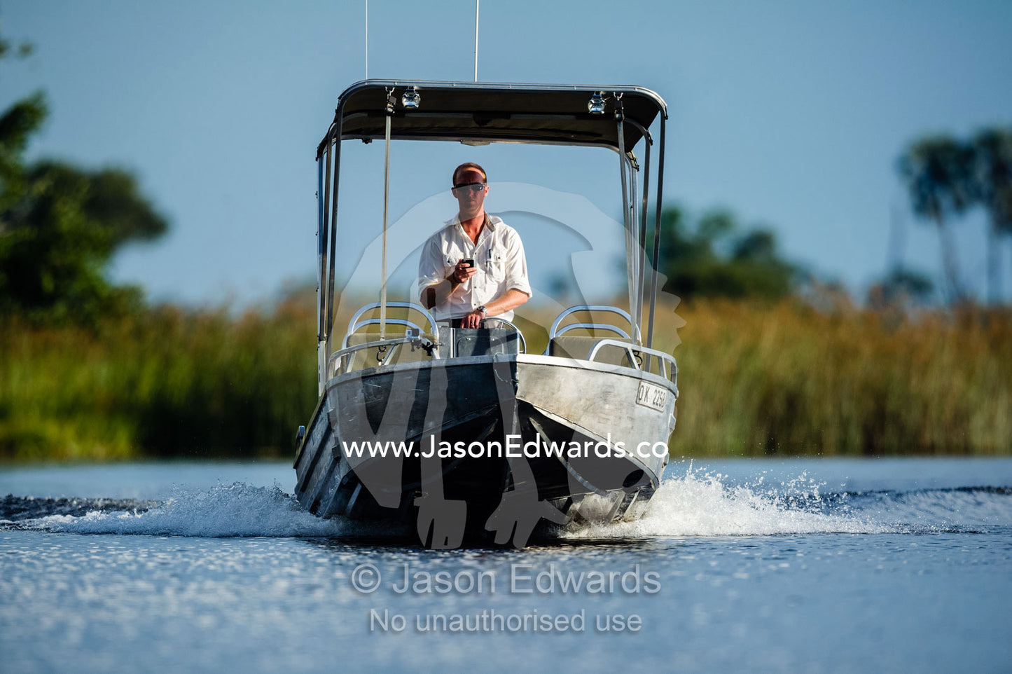 A safari guide texting on a cell phone whilst driving a boat on a river. Selinda Spillway, Selinda Reserve, Linyanti Region, Northern Botswana.