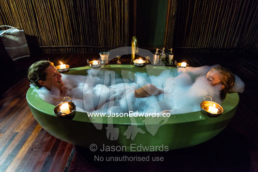 A mother and son soaking in a bubble bath in a luxury safari camp tent with candles. Selinda Camp, Selinda Reserve, Linyanti Region, Northern Botswana.