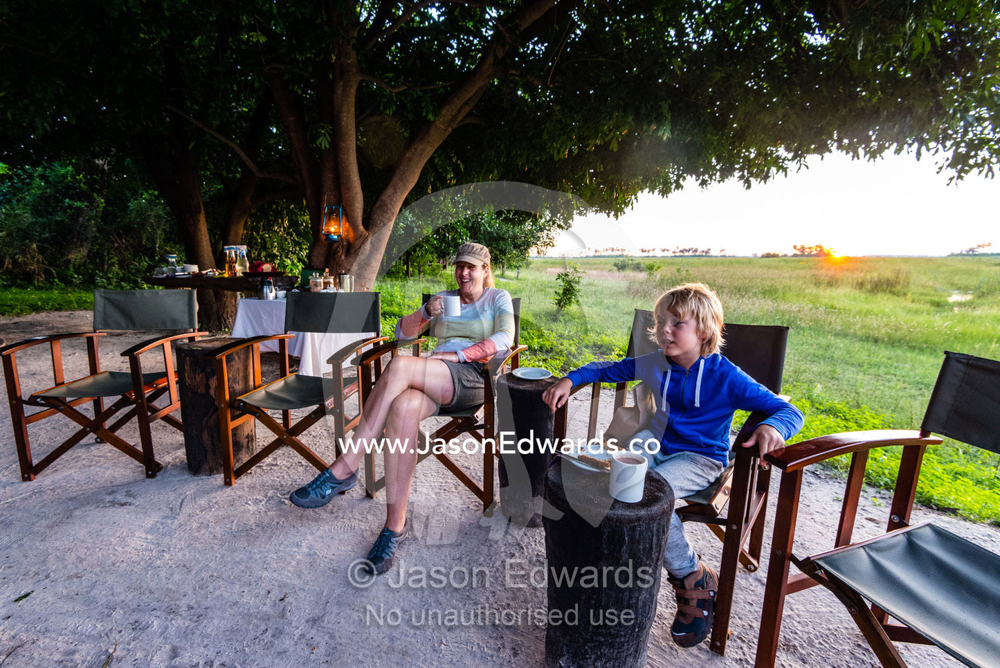 A mother and son sitting around a campfire at dawn in a luxury safari camp. Selinda Camp, Selinda Reserve, Linyanti Region, Northern Botswana.