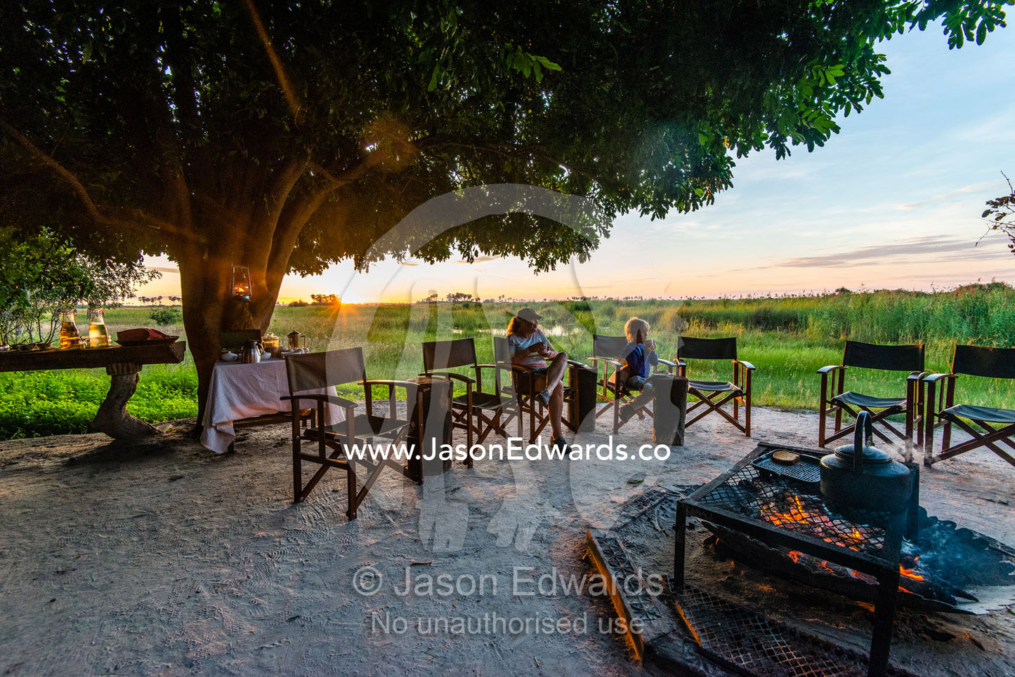 A mother and son sitting around a campfire at dawn in a luxury safari camp. Selinda Camp, Selinda Reserve, Linyanti Region, Northern Botswana.