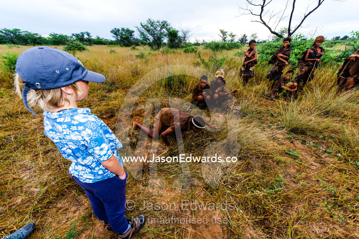 A boy watches a San Bushmen demonstrate hunting for antelope. Kalahari Desert, Botswana.