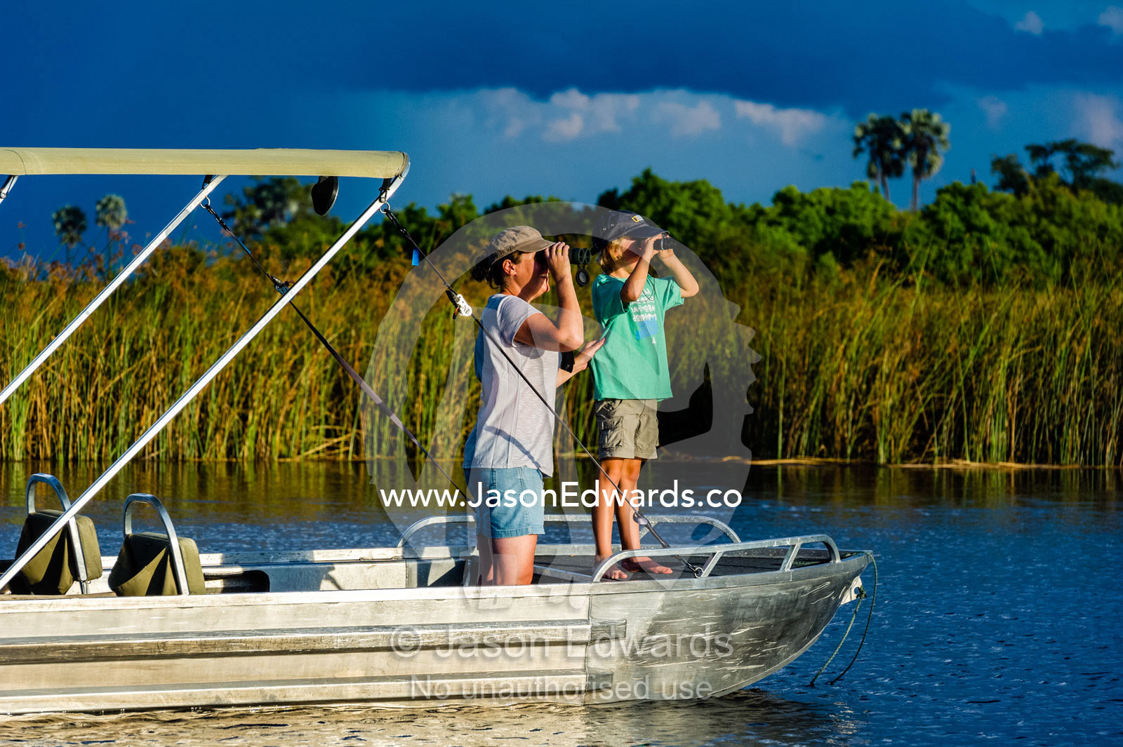 A mother and son watching river wildlife from a safari boat at sunset. Selinda Spillway, Selinda Reserve, Linyanti Region, Northern Botswana.