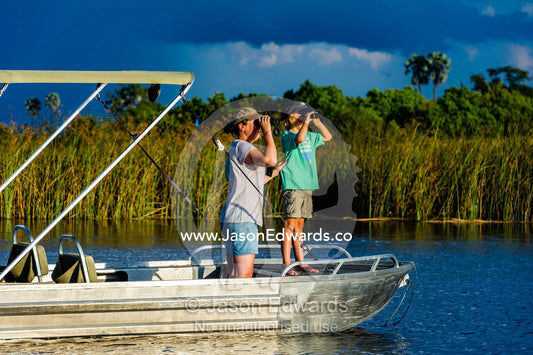 A mother and son watching river wildlife from a safari boat at sunset. Selinda Spillway, Selinda Reserve, Linyanti Region, Northern Botswana.