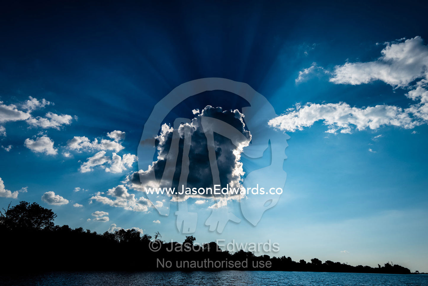God rays pass from behind clouds in a clear blue sky above a river. Chobe River, Chobe National Park, Botswana.