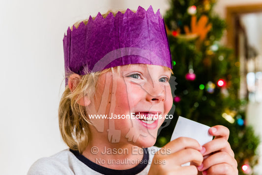A boy wears a paper hat and reads a joke from a bon bon on Christmas Day. Carlton North, Melbourne, Victoria, Australia.
