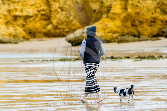 A woman walking a terrier dog along a beach at low tide in winter. Torquay, Surf Coast, Victoria, Australia.