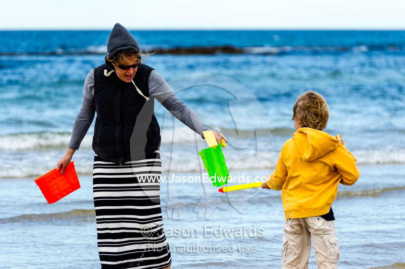 A boy squirts his mother with a water pistol on a beach in summer. Torquay, Surf Coast, Victoria, Australia.