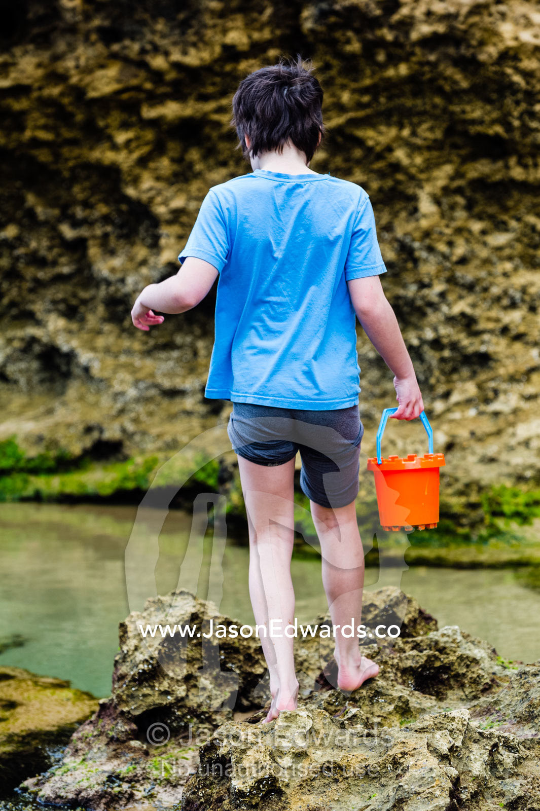 A girl carries a sand castle bucket across rocks on a beach. Torquay, Surf Coast, Victoria, Australia.