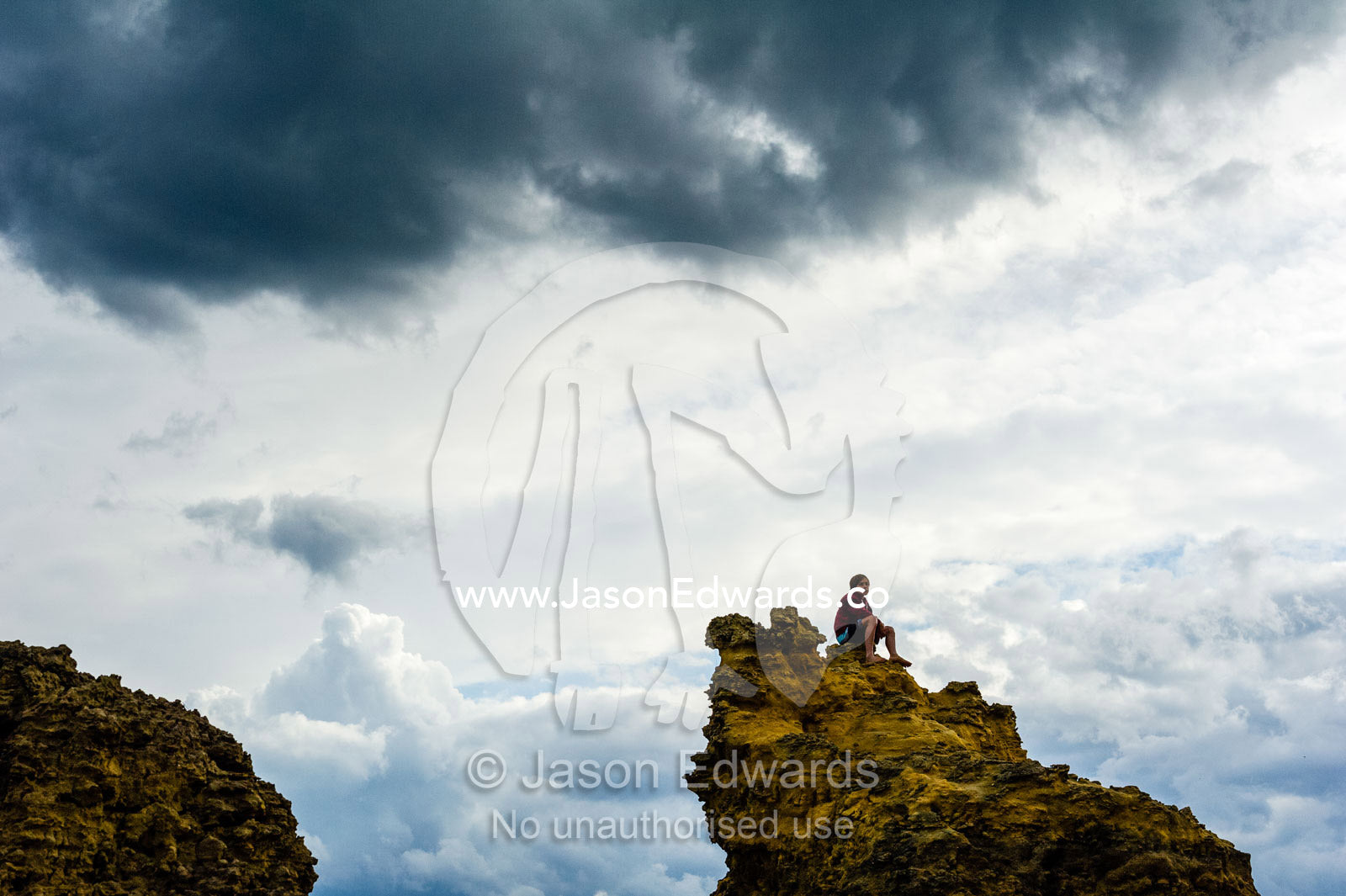 The silhouette of a boy sitting on top of a jagged rock on a beach. Torquay, Surf Coast, Victoria, Australia.