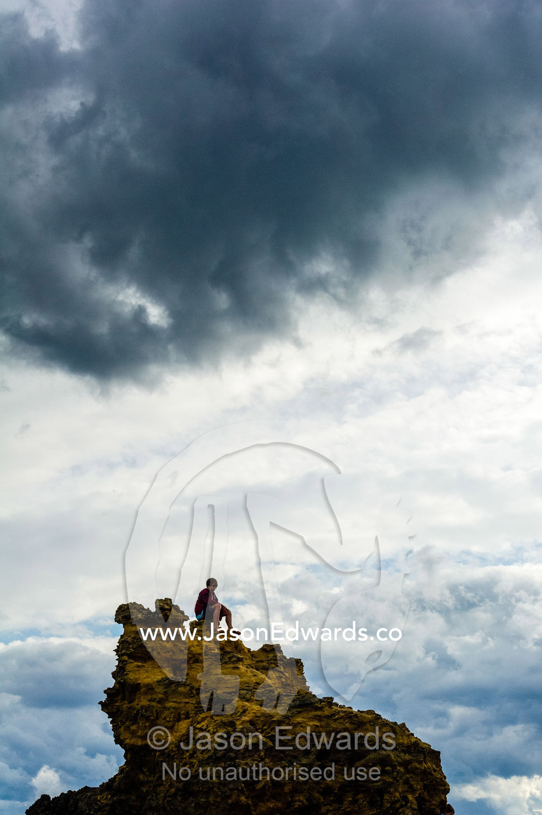 The silhouette of a boy sitting on top of a jagged rock on a beach. Torquay, Surf Coast, Victoria, Australia.
