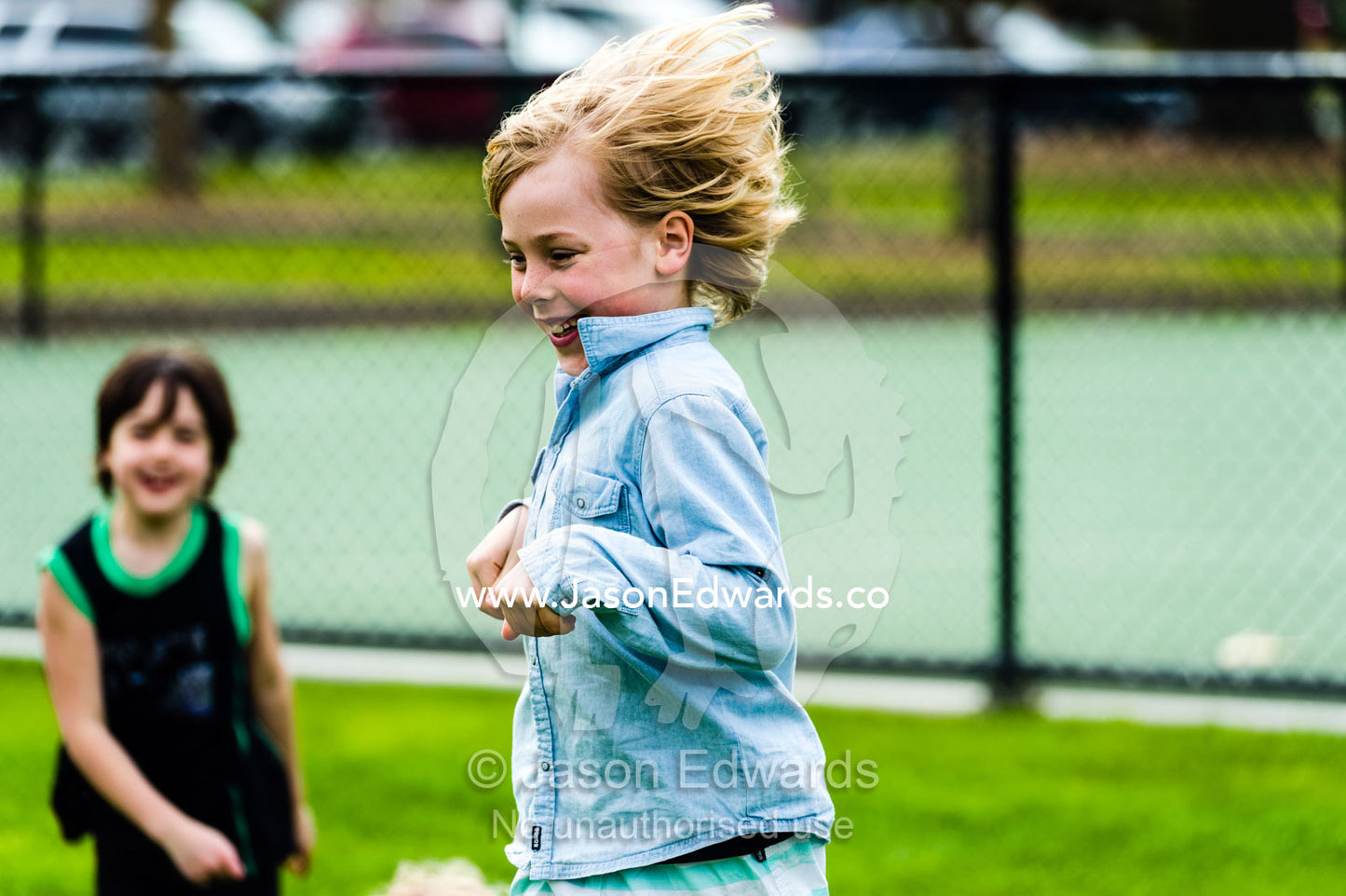 A boy hops like a kangaroo on a sports field in front of his friend. Albert Park, Melbourne, Victoria, Australia.
