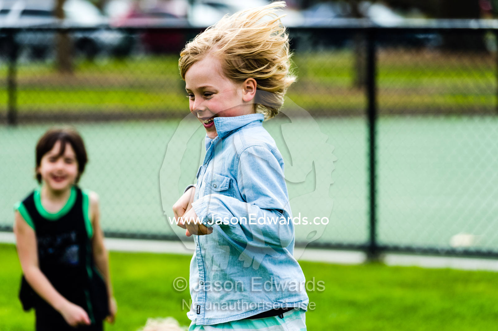 A boy hops like a kangaroo on a sports field in front of his friend. Albert Park, Melbourne, Victoria, Australia.