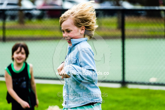 A boy hops like a kangaroo on a sports field in front of his friend. Albert Park, Melbourne, Victoria, Australia.