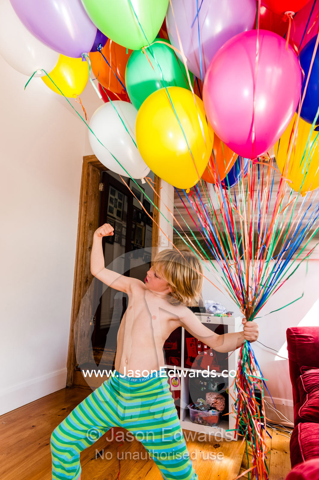 A boy displays his muscles whilst holding a bunch of colorful helium birthday balloons. Carlton North, Melbourne, Victoria, Australia.