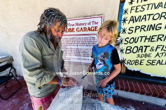 An artist helps a boy carve a shark into a block of sandstone during a school holiday program activity. Robe, Limestone Coast, South Australia, Australia.