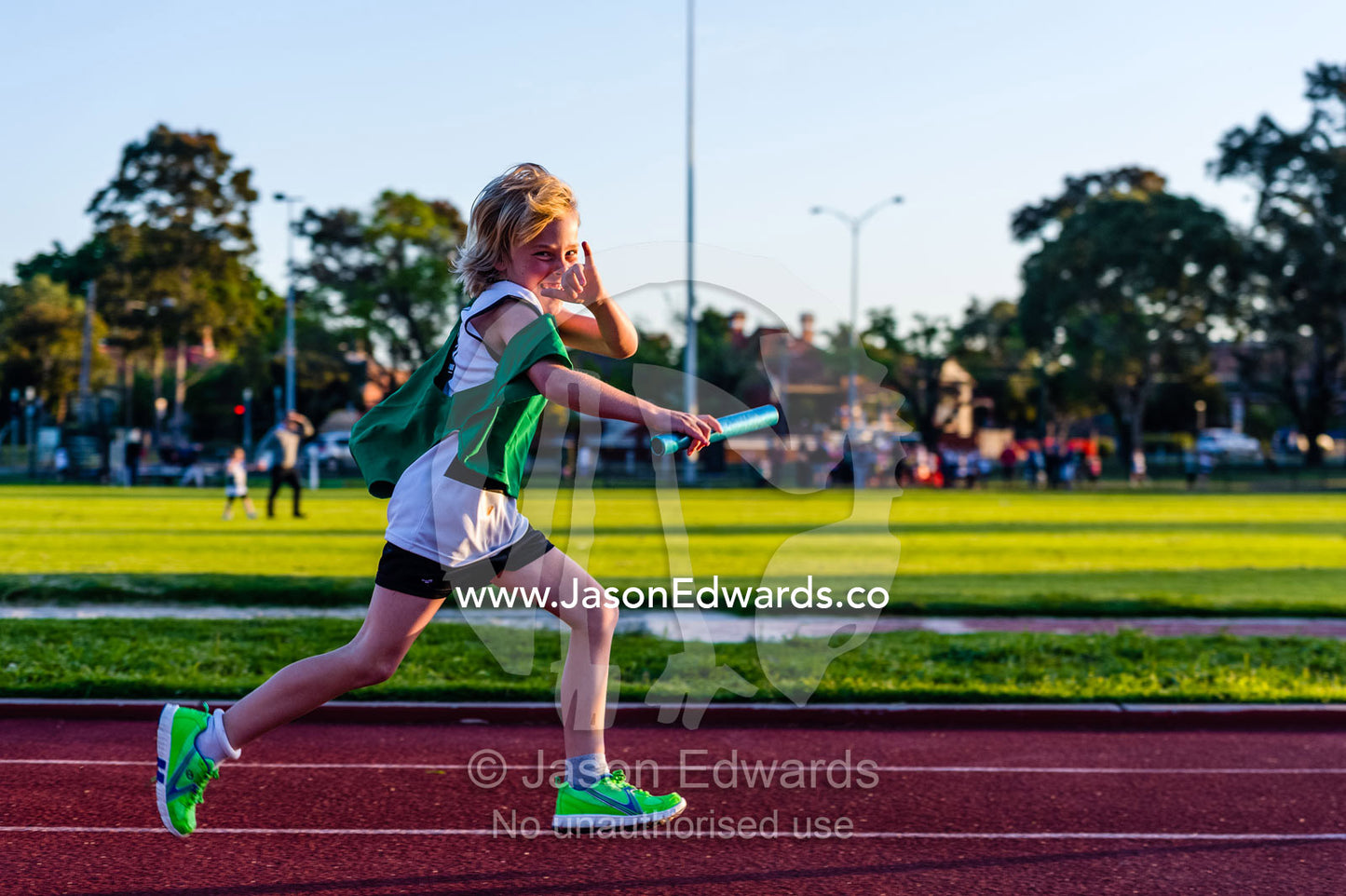 A laughing boy runs a baton relay on an athletics track. Collingwood, Melbourne, Victoria, Australia.