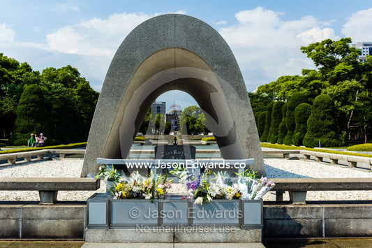 A saddle-shaped monument covers a cenotaph holding the names of all of the people killed by the bomb. The monument frames the Peace Flame and the A-Bomb Dome. Hiroshima Peace Memorial Park, Hiroshima, Honshu Island, Japan.