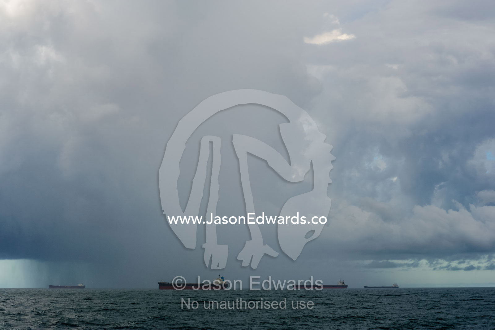A menacing tropical rainstorm darkens the ocean and sky over cargo ships at anchor. Gladstone, Queensland, Australia.