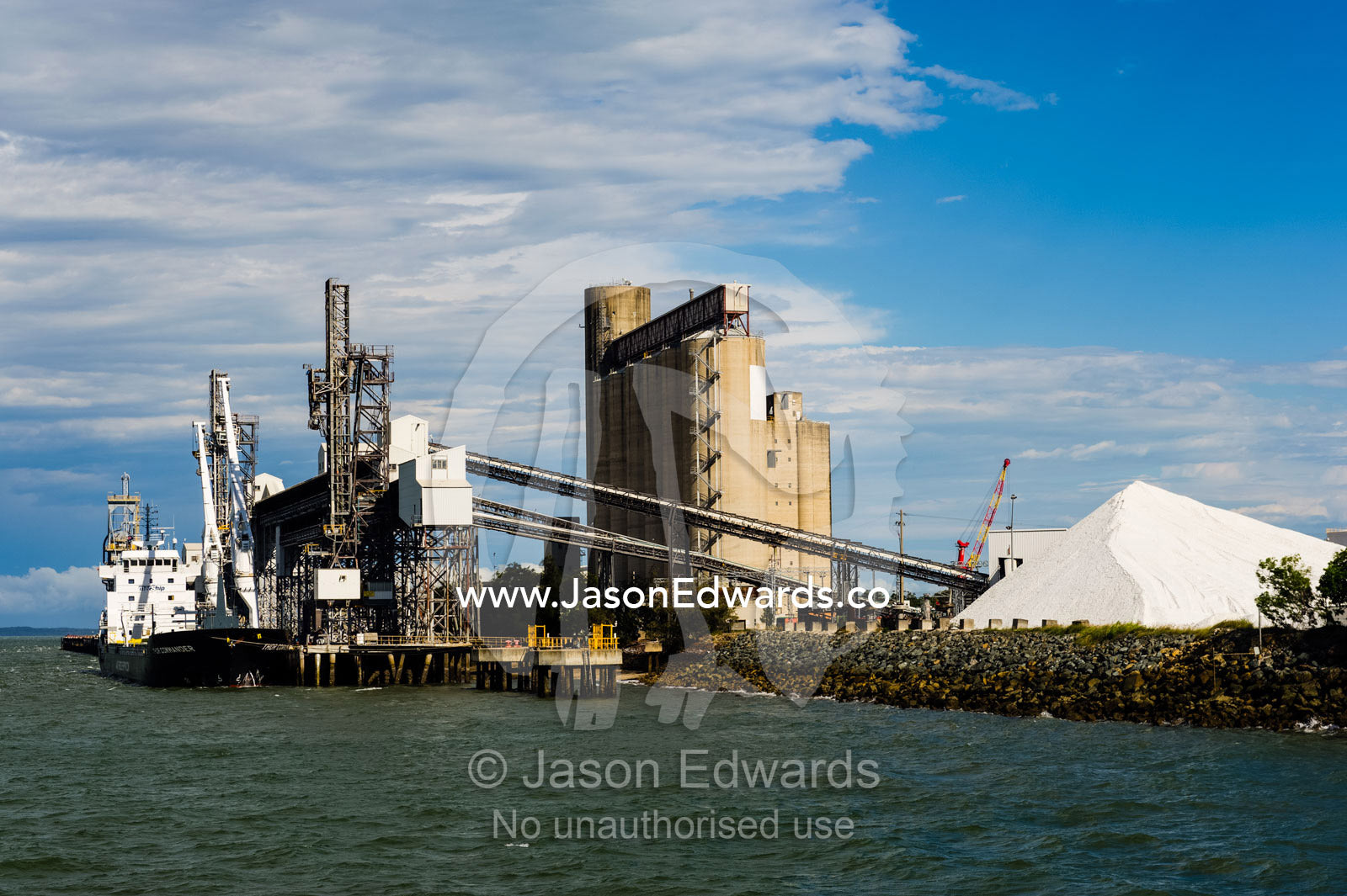A project type ship loading wheat or bulk cargo. The stockpile of white material is calcite, used in industrial manufacturing, including toothpaste. Gladstone, Queensland, Australia.