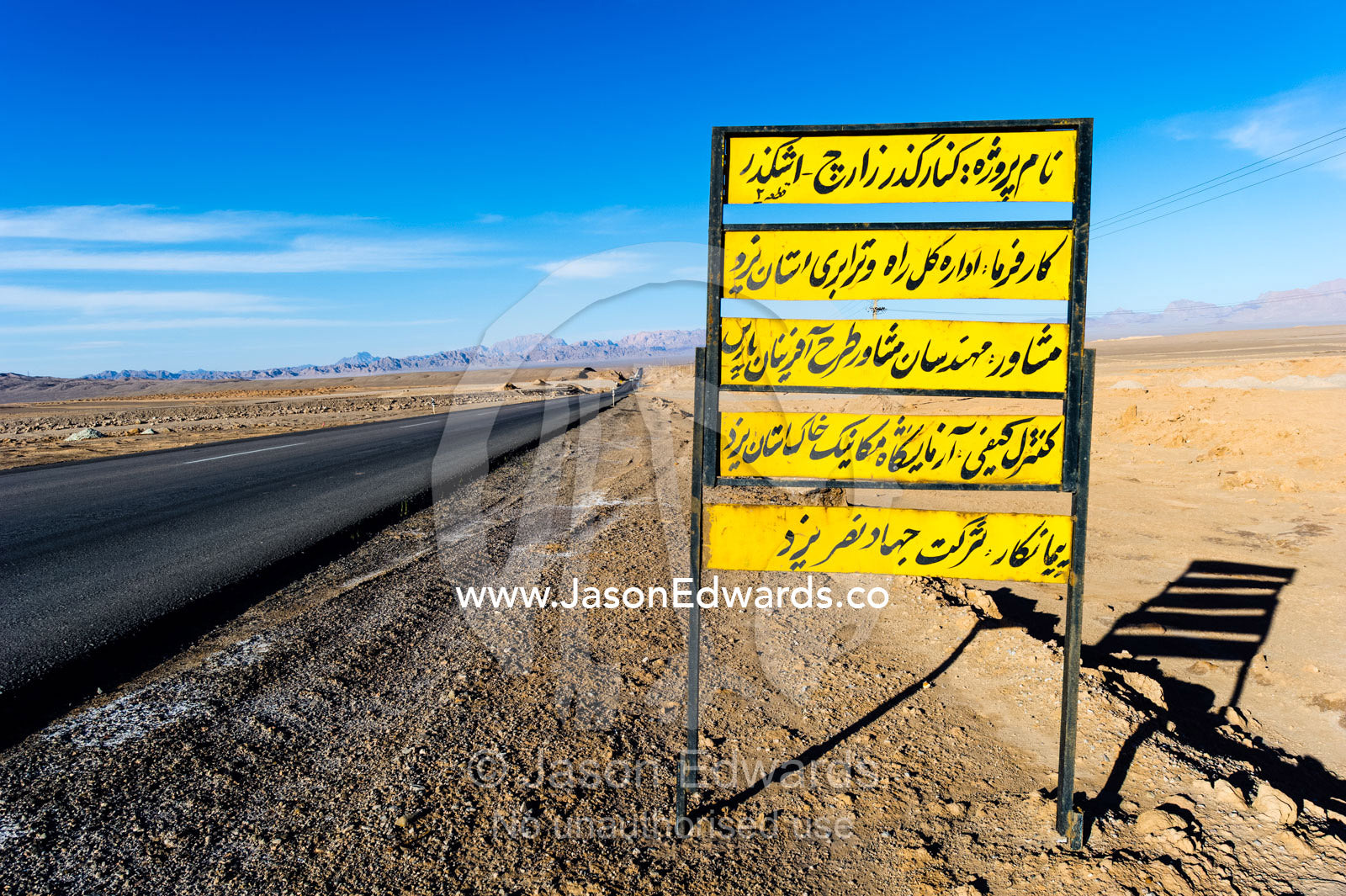 A road sign in Arabic beside a long straight road crosses the desert towards the mountains. Kharanagh, Yazd, Yazd Province, Islamic Republic of Iran.