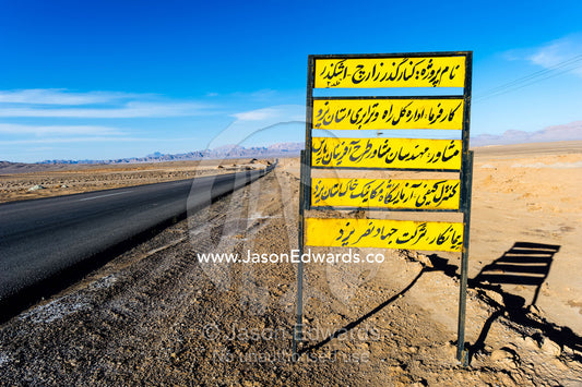 A road sign in Arabic beside a long straight road crosses the desert towards the mountains. Kharanagh, Yazd, Yazd Province, Islamic Republic of Iran.