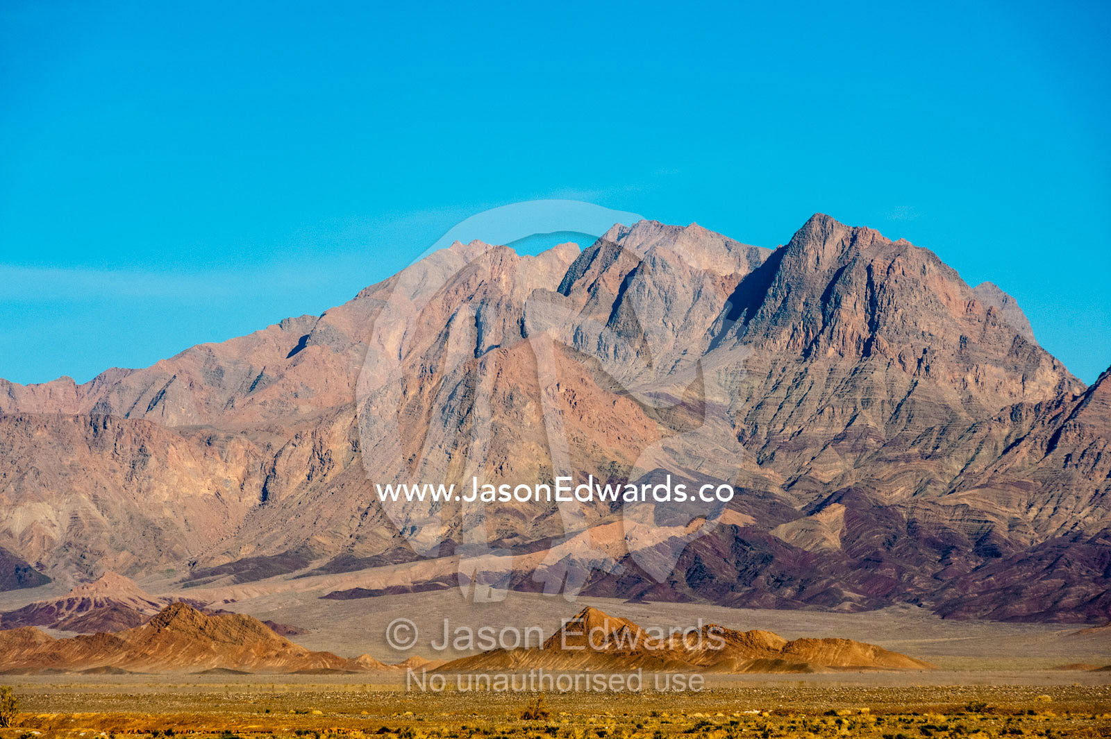 Erosion on the slopes of jagged desert mountain peaks beneath a clear blue sky. Kharanagh, Yazd, Yazd Province, Islamic Republic of Iran.