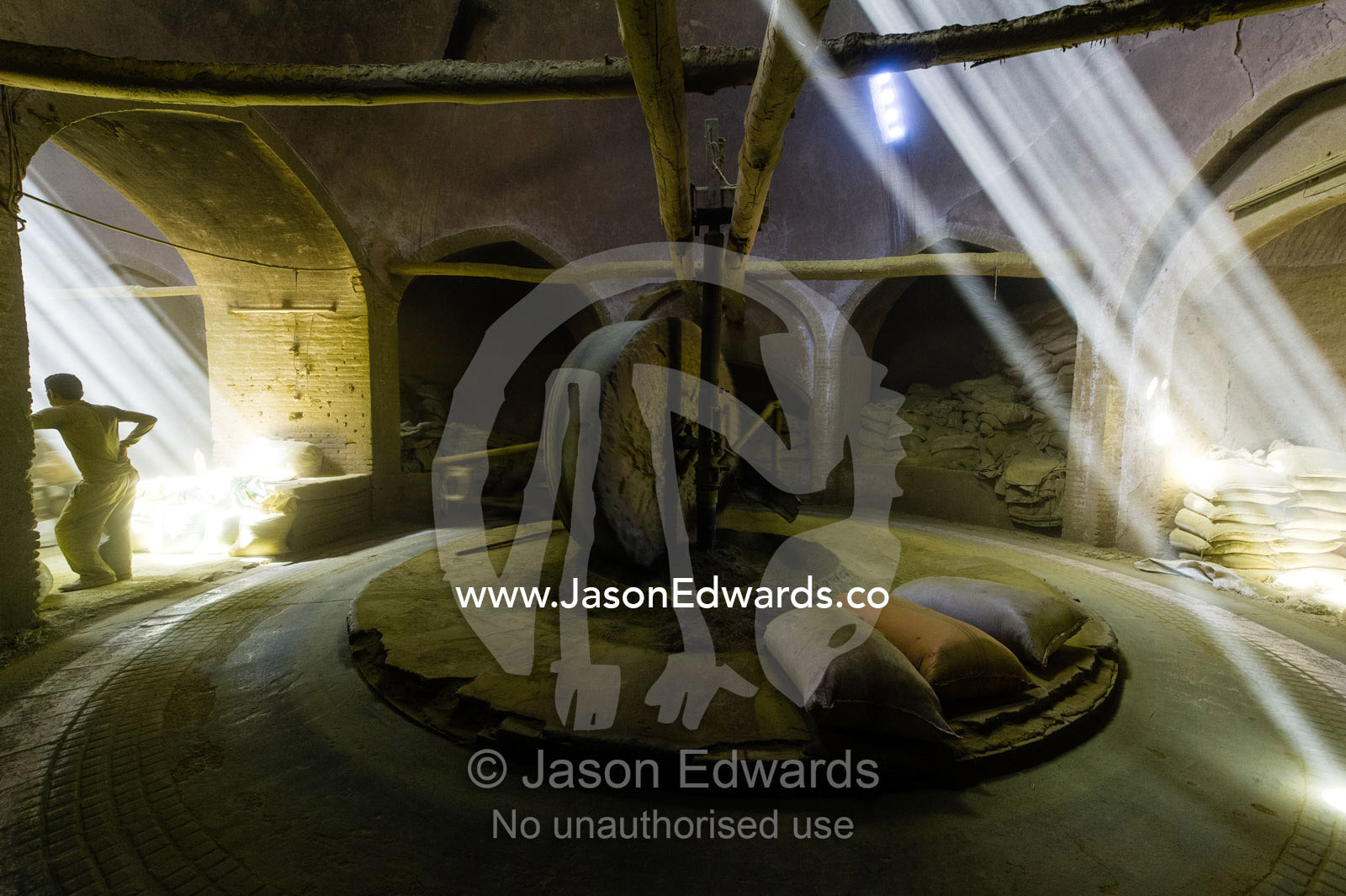 A labourer rests whilst sunrays light a giant stone wheel grinding henna seeds into powder in an ancient henna mill. Yazd, Yazd Province, Islamic Republic of Iran.