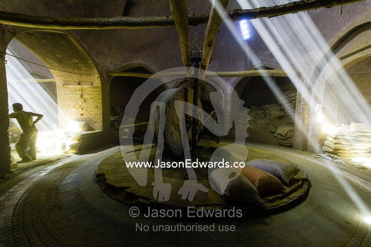 A labourer rests whilst sunrays light a giant stone wheel grinding henna seeds into powder in an ancient henna mill. Yazd, Yazd Province, Islamic Republic of Iran.