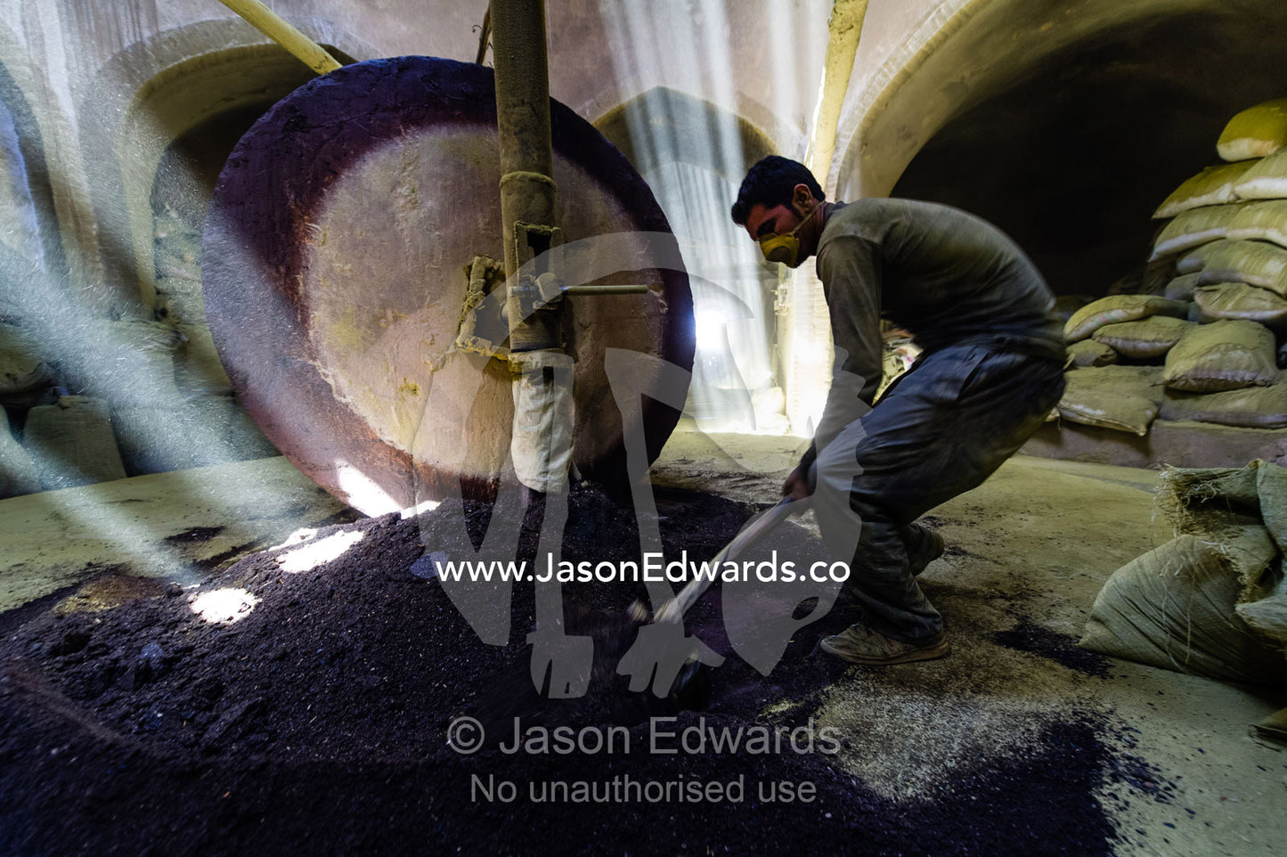 A labourer shovels henna seeds in front of a stone grinding wheel in a henna mill. Yazd, Yazd Province, Islamic Republic of Iran.