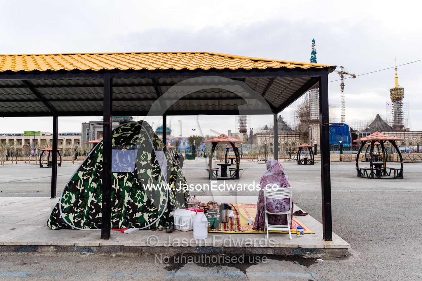 A woman camped beneath a bus shelter beside a mausoleum. Mausoleum of Ayatollah Khomeini, Tehran, Tehran Province, Islamic Republic of Iran.
